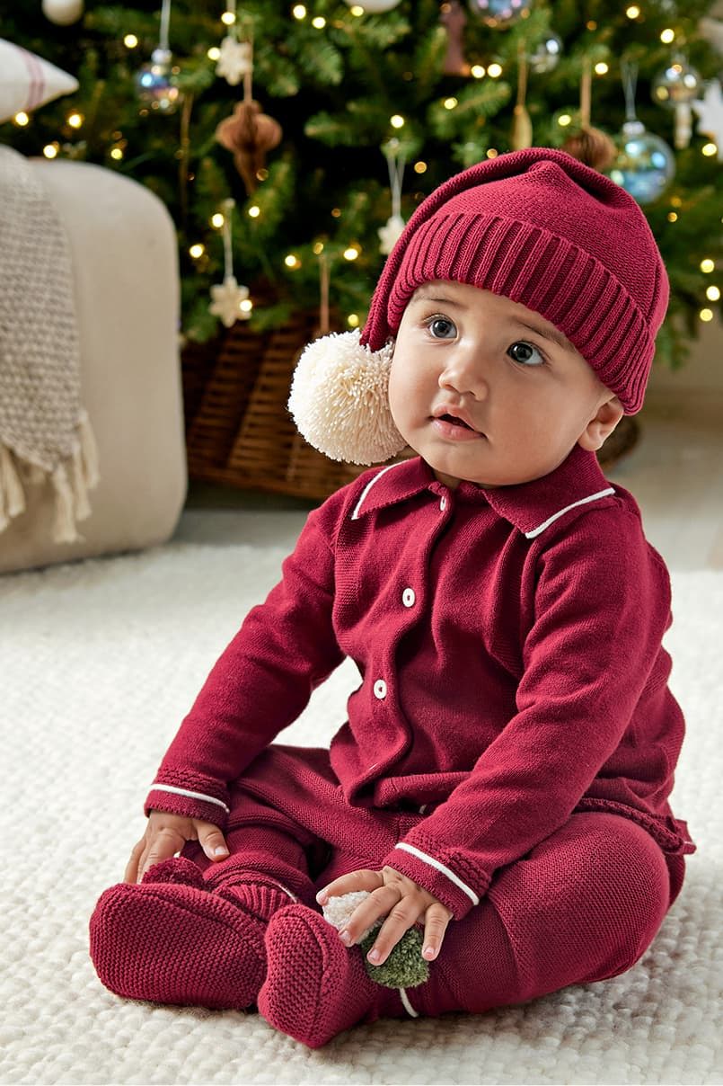 Baby in red outfit and hat sitting by a Christmas tree.