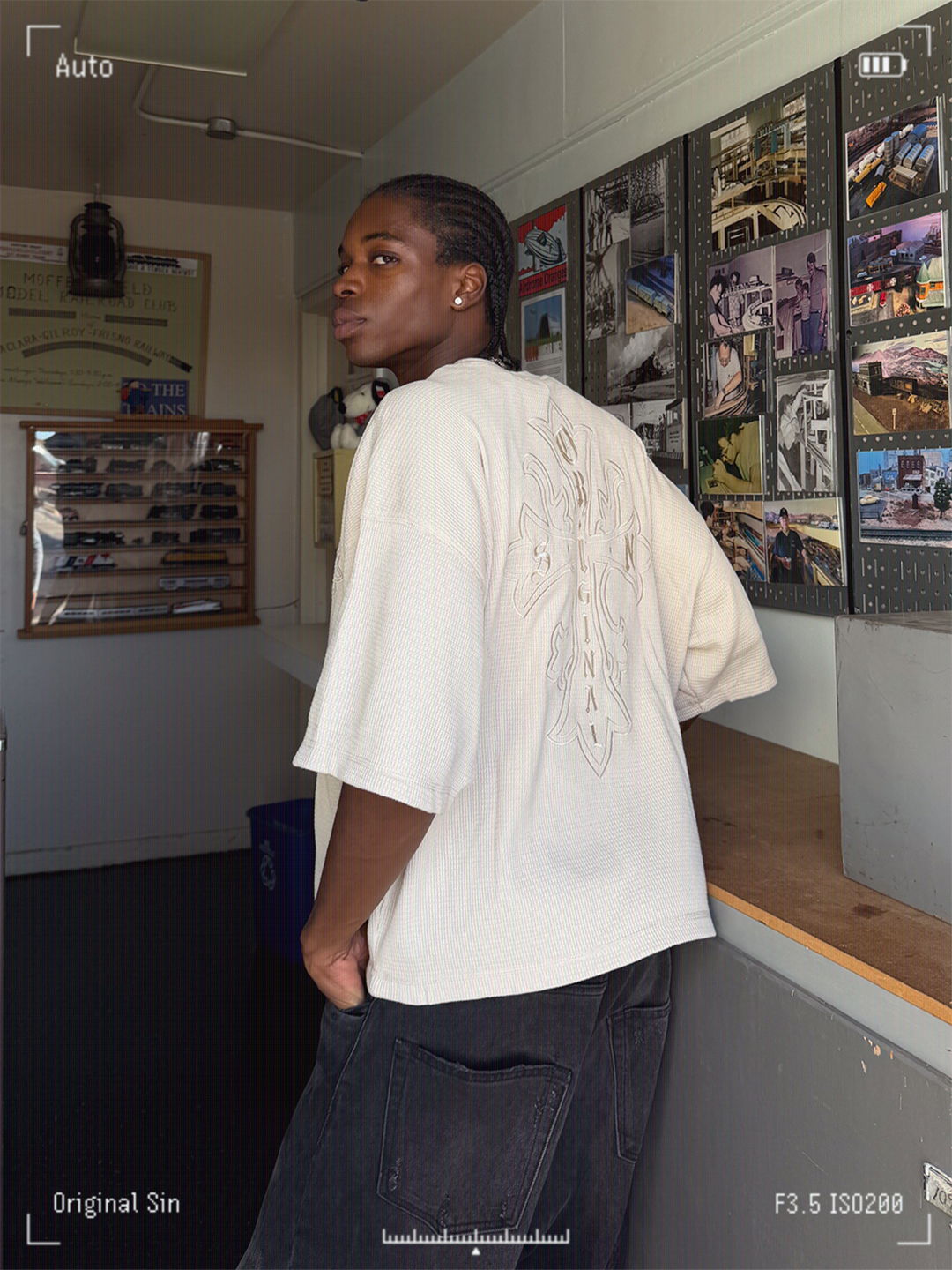 Person in a beige shirt posing in a room with photographs on the wall.