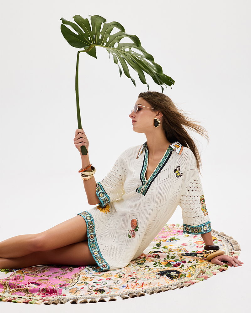 Woman in a white dress holding a palm leaf, sitting on a colorful rug.