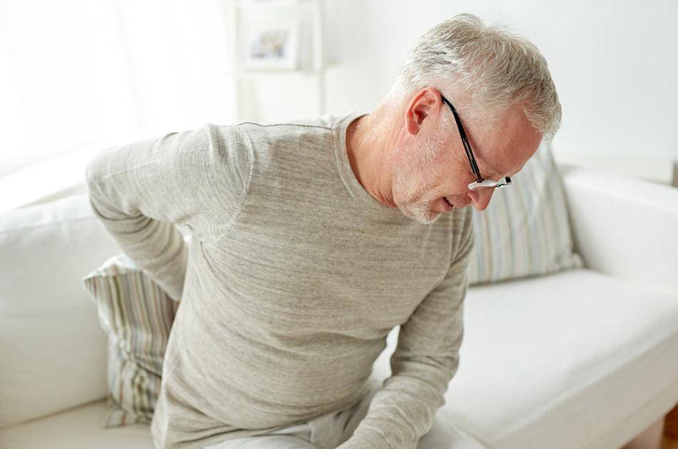 Elderly man clutching his lower back with his right hand while sitting on a white sofa