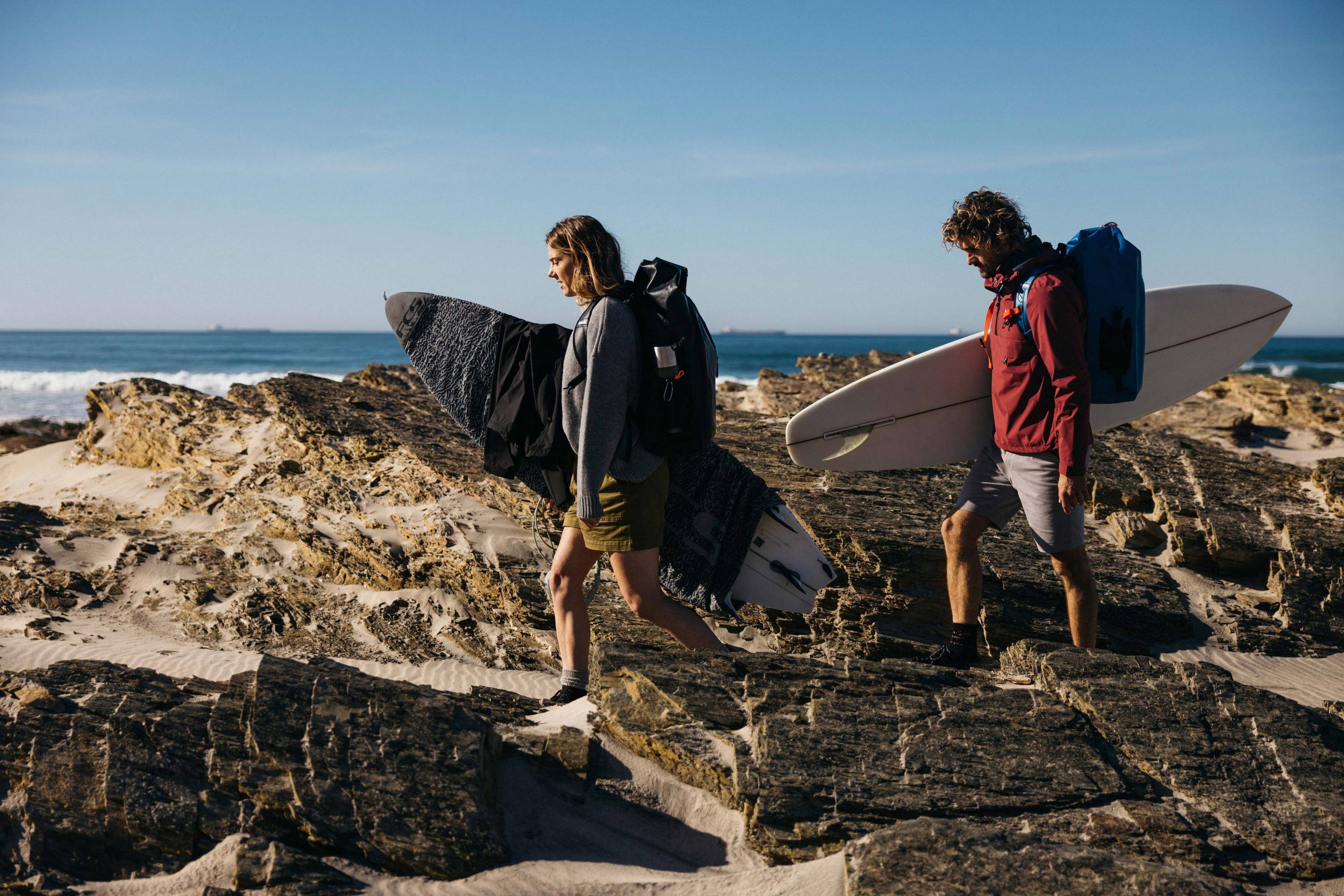 Lucy and Alex carry their surfboards over rocks in Portugal