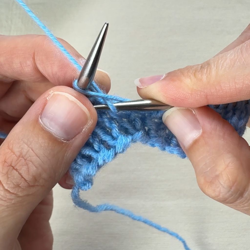 Detailfoto, wie eine rechte Masche gestrickt wird. Der Faden wird durch die Masche durchgezogen.