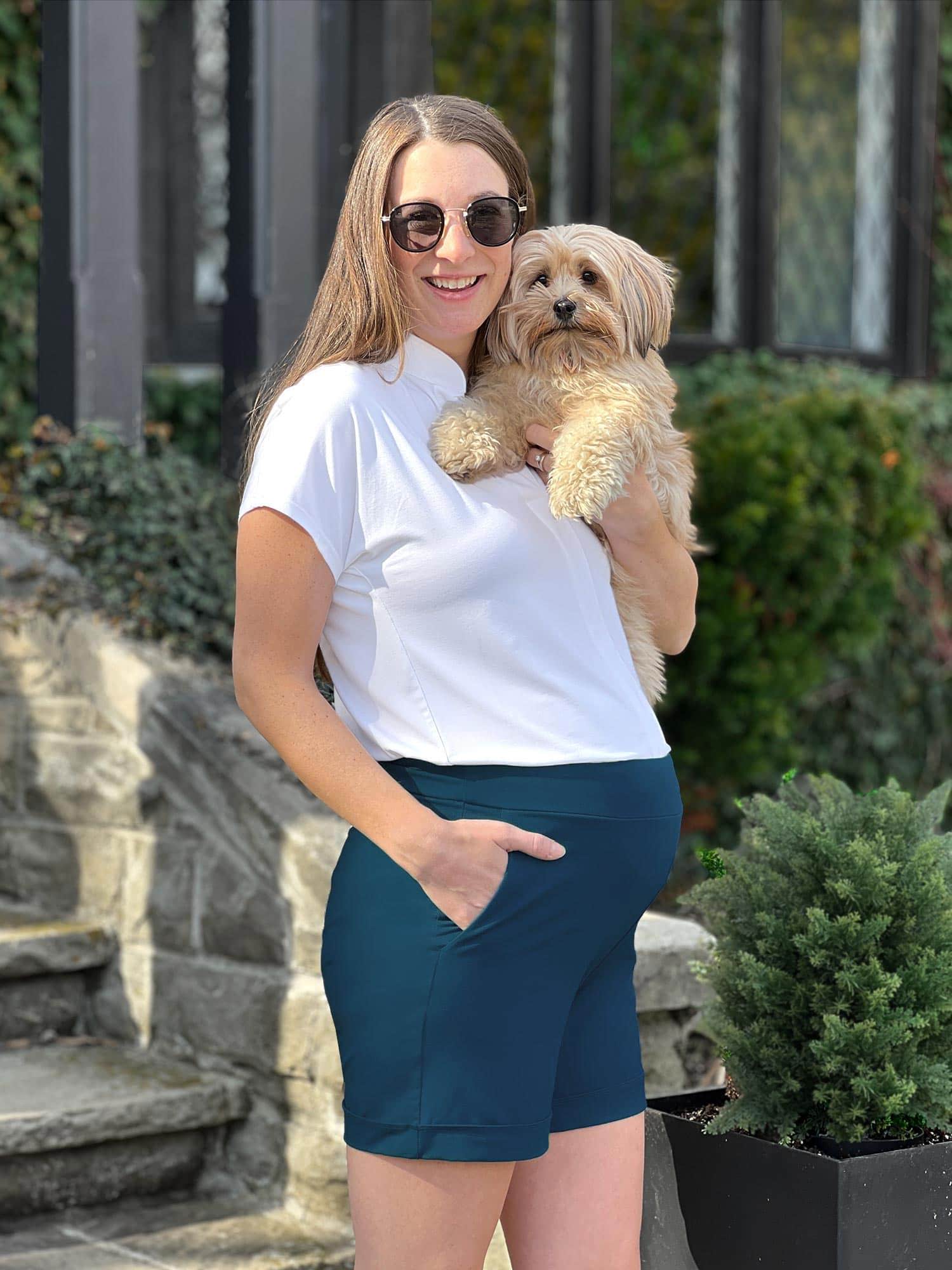 Pregnant woman standing wearing Miik's Leland everyday dressy short in navy with a white top and holding a dog.