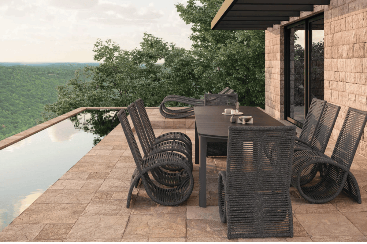 Outdoor dining area with a dark rectangular dining table and sculptural woven dining chairs beside an infinity pool overlooking a wooded landscape.