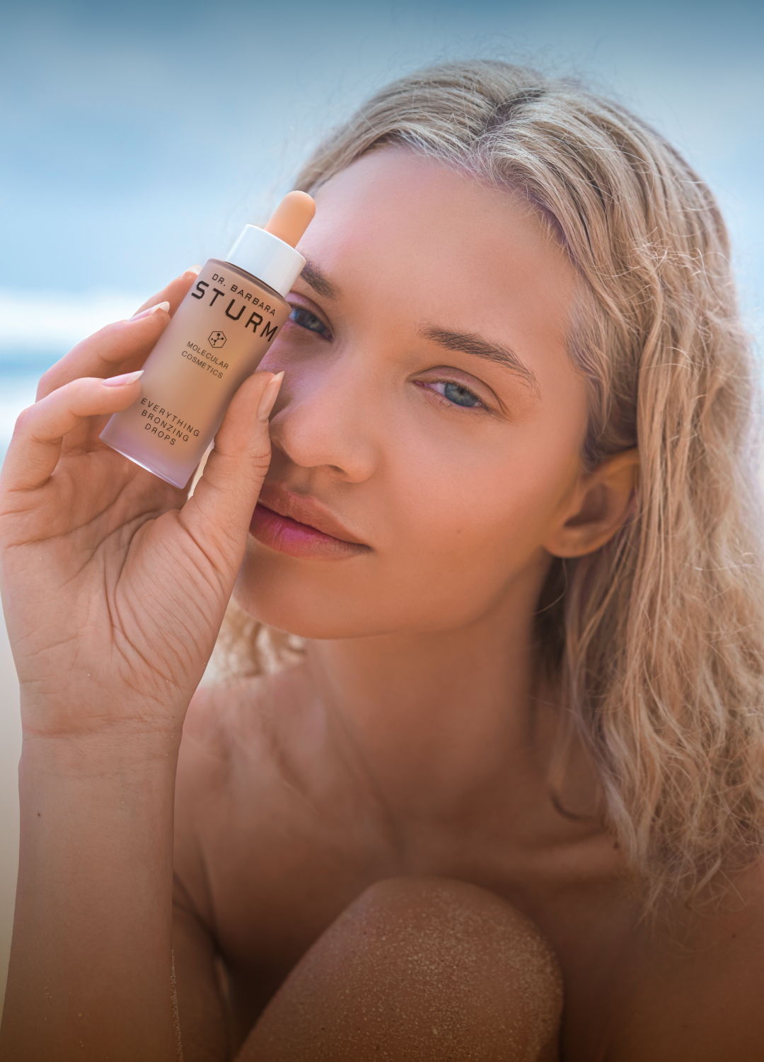 Woman holding skincare product near her face on a beach.