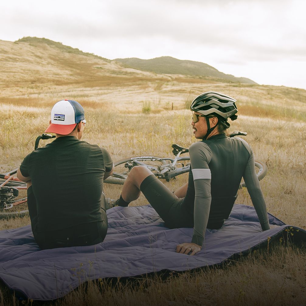Two cyclists sit on a blanket in a grassy landscape, enjoying the view.
