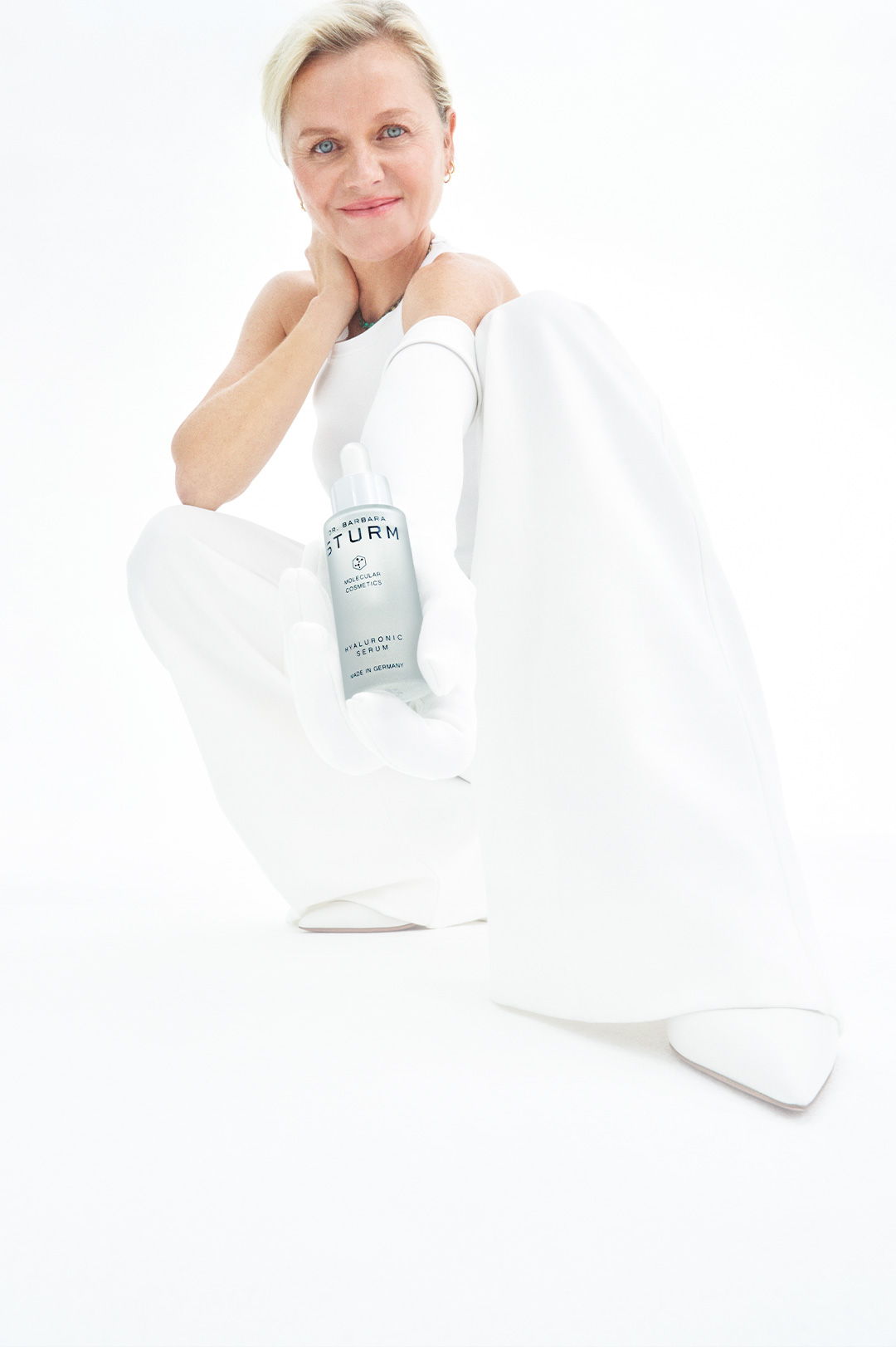 Woman in white outfit holding a bottle, smiling against a white background.