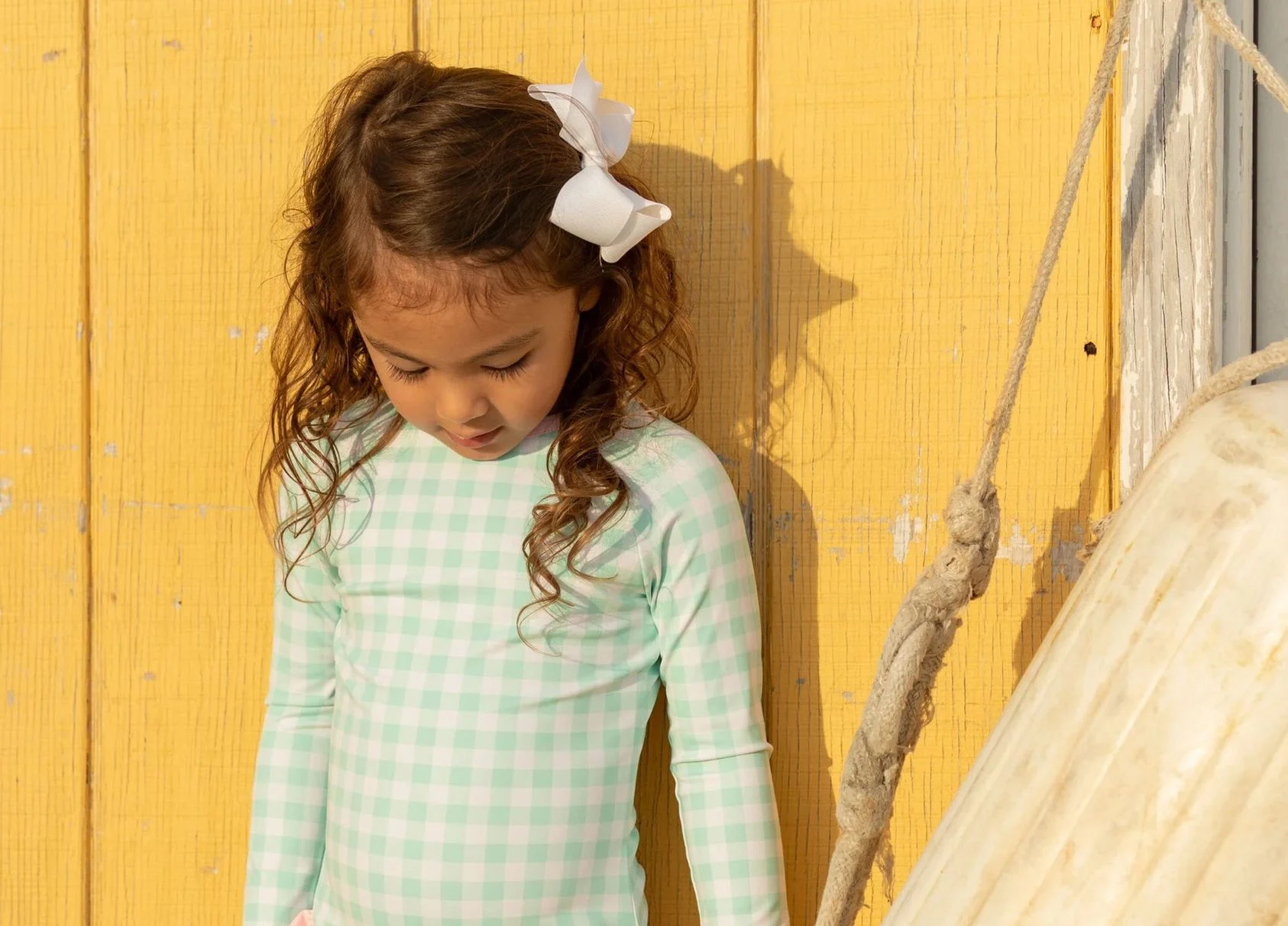 Girl with curly hair in a swimsuit standing by a yellow wall.
