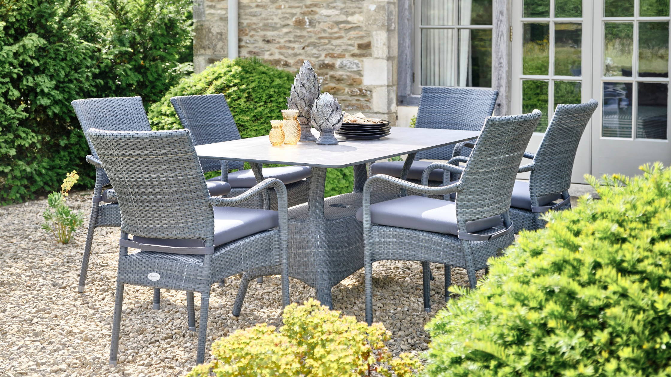 Grey rattan dining set on a shingle patio surrounded by hedges.