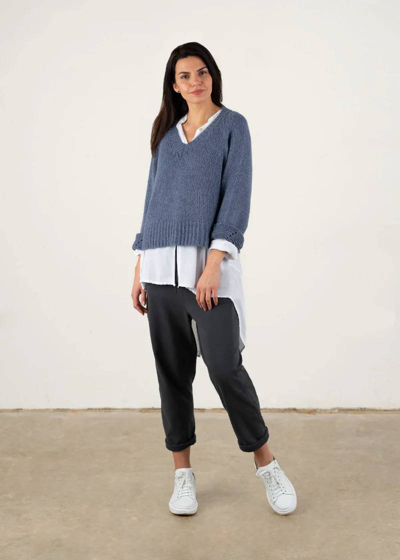 A model wearing a grey blue knitted sweater ove a white shirt, grey trousers and white trainers