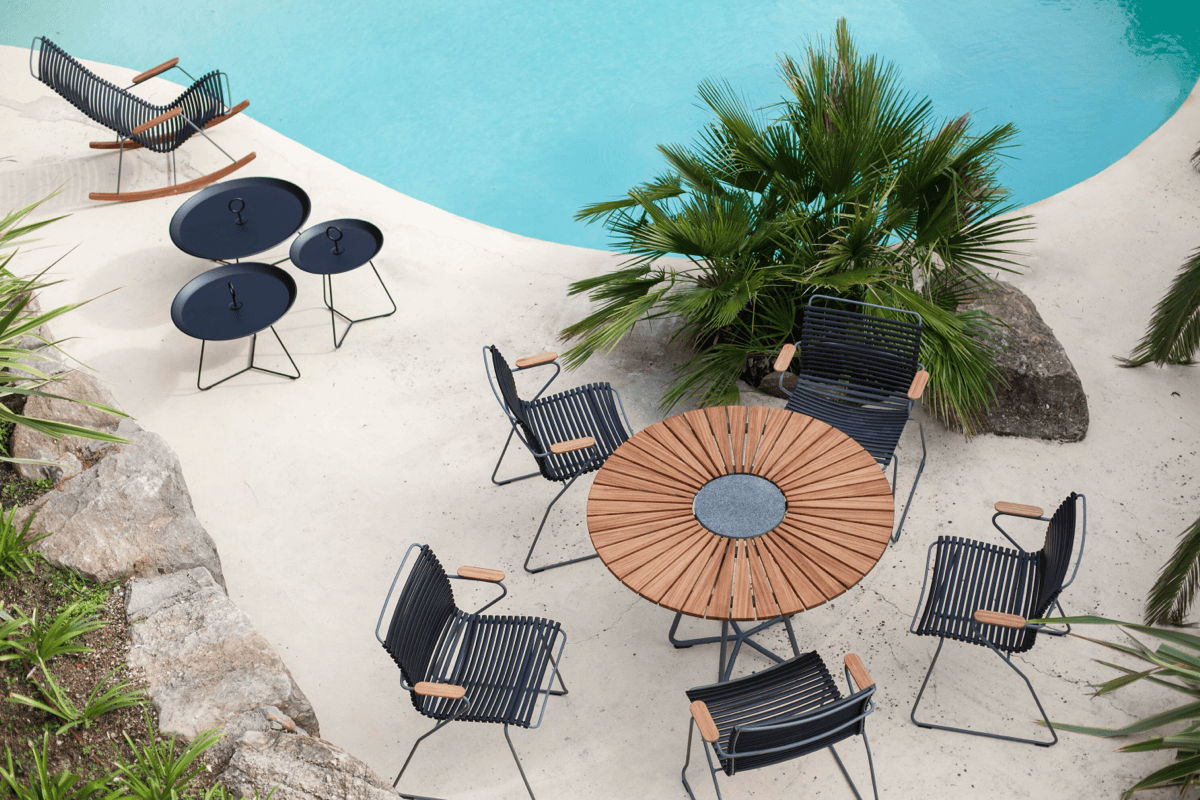 Outdoor dining set with a round slatted wood table and black chairs on a sandy poolside patio with palm trees.