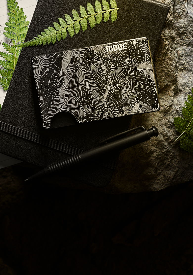 A Ridge wallet on a notebook with a pen and ferns.