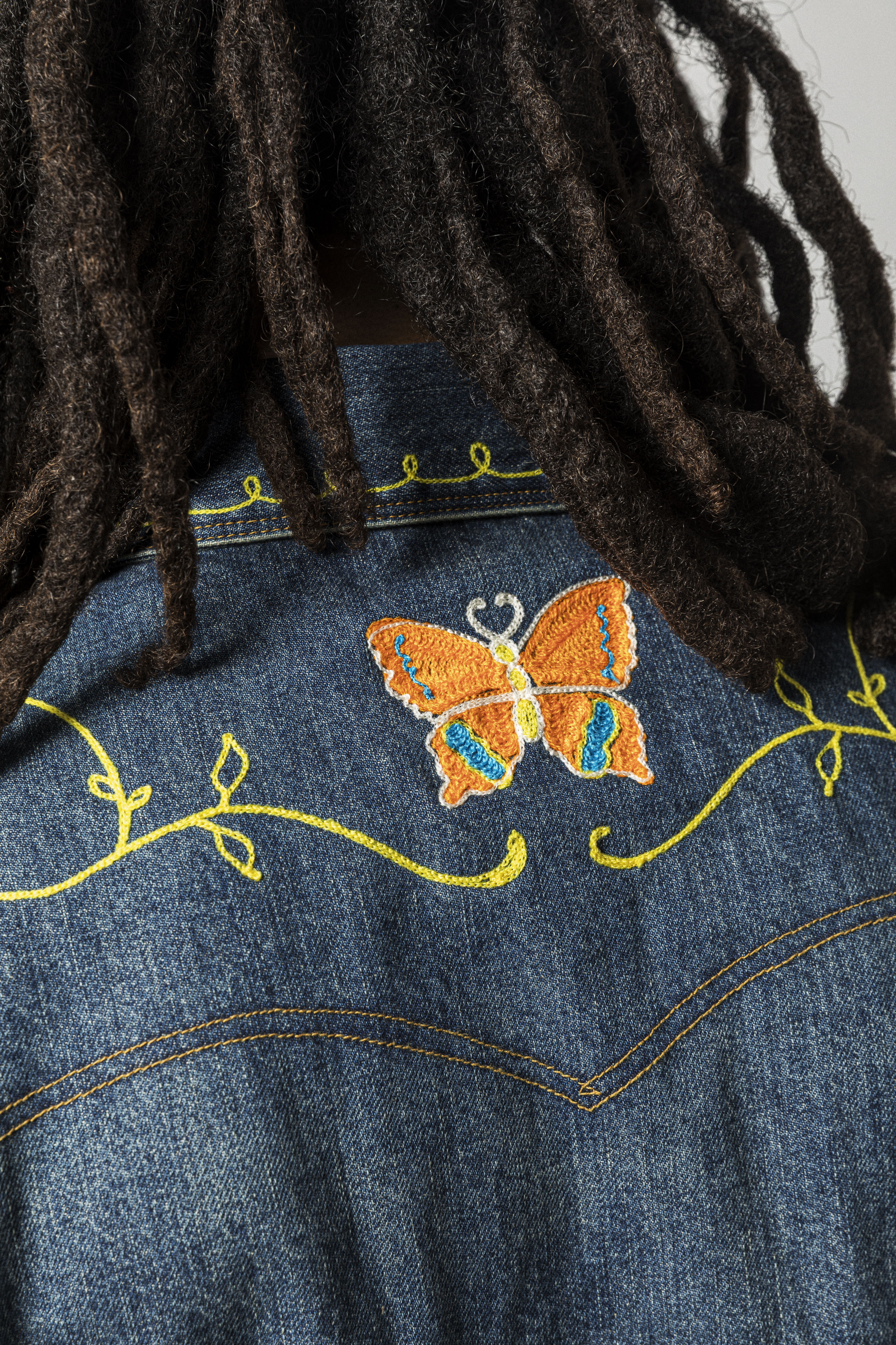 Person with dreadlocks wearing a denim jacket featuring butterfly embroidery on the back.