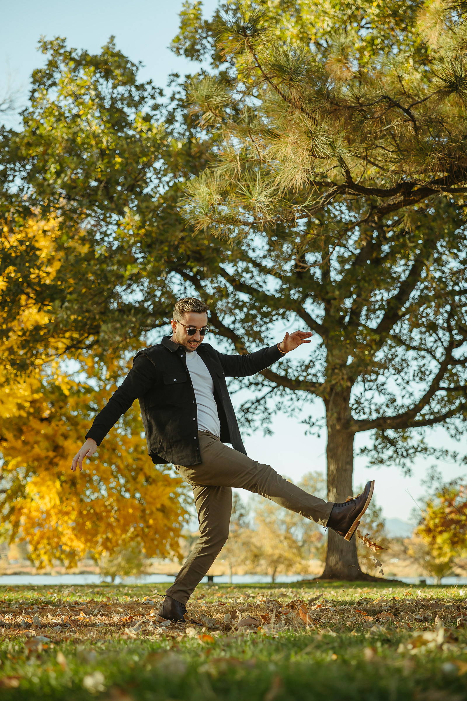 Man dancing in the autumn leaves wearing brown boots, white t shirt, a black overshirt, and olive chinos from Under510.com