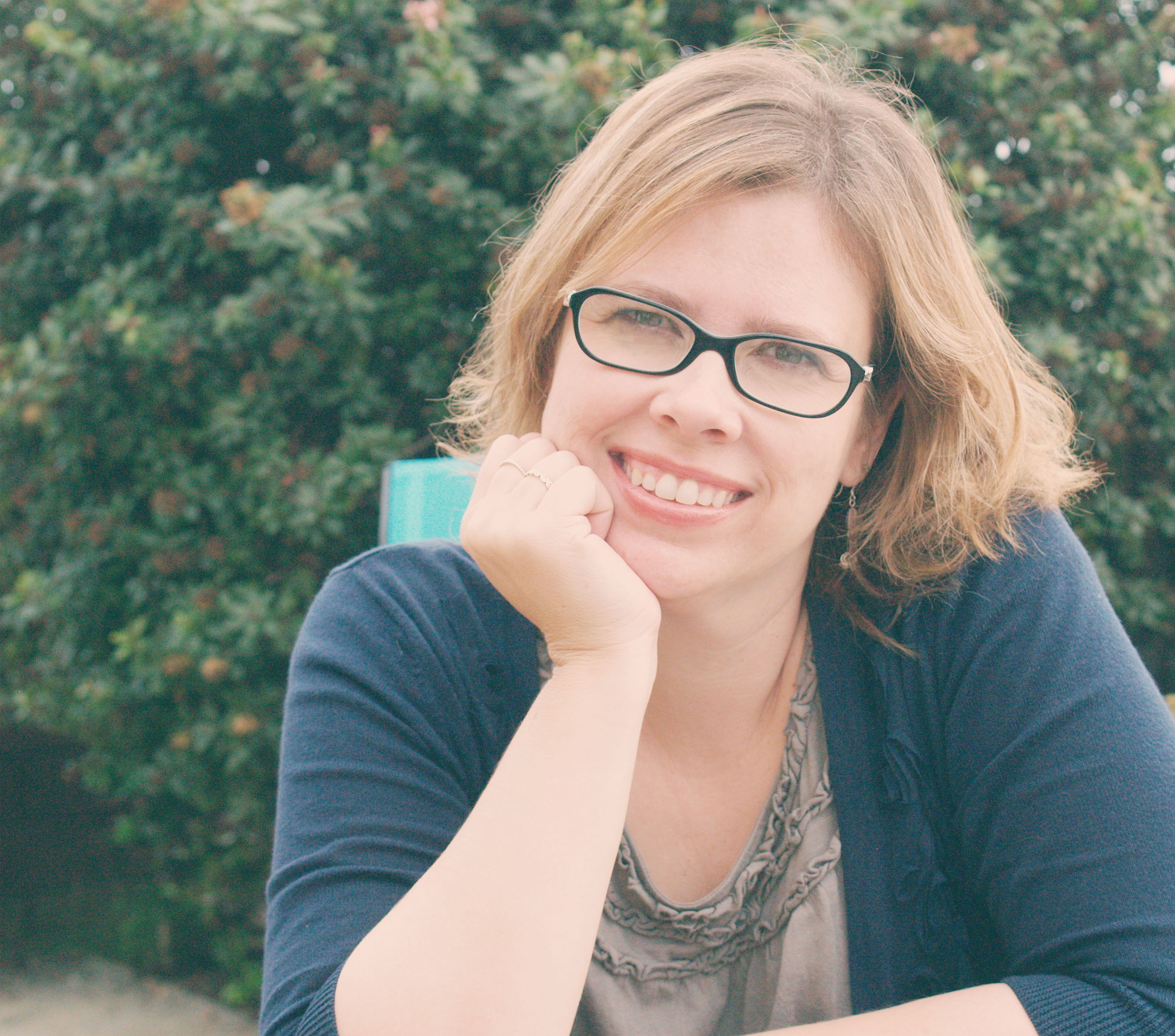 Woman with glasses smiling, resting chin on hand, surrounded by greenery.