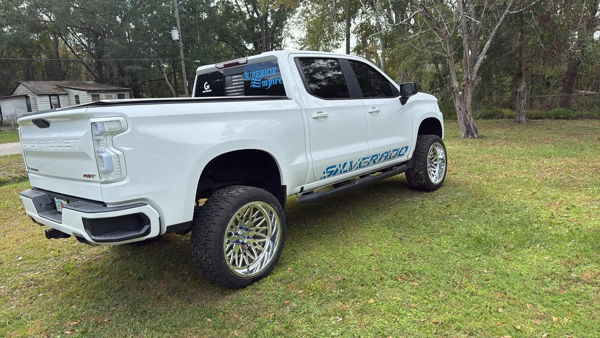 Rear three-quarter Joel's lifted white Chevy Silverado RST parked on grass with blue decals photo 1
