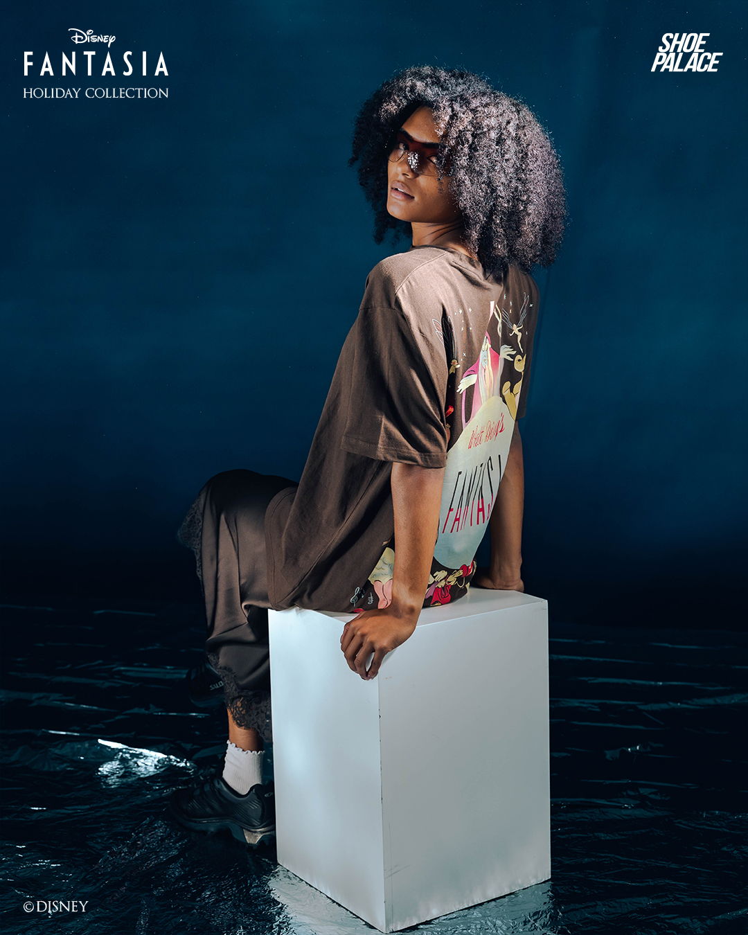 Person with curly hair sitting on a white cube against a blue background.