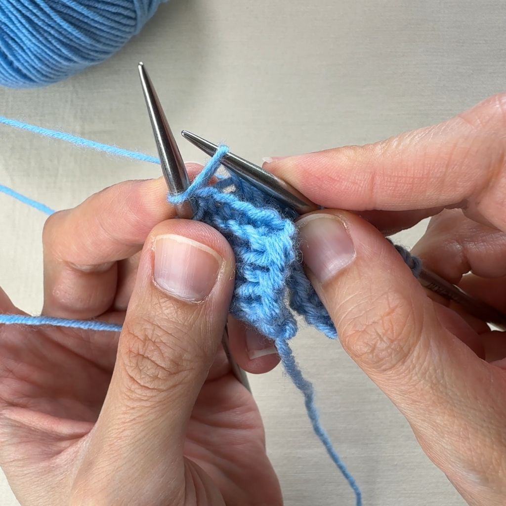 Detailfoto, wie eine rechte Masche gestrickt wird. Die Nadel sticht von unten in die Masche ein, der Faden liegt hinter der Arbeit.