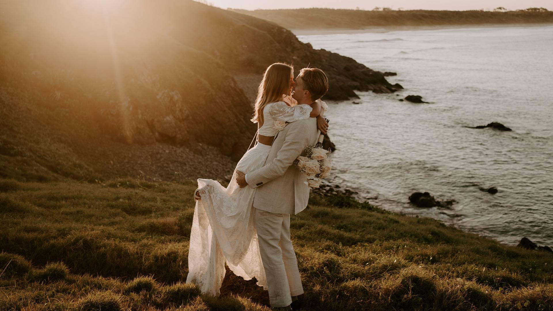 Groom carrying his bride on the coastline wearing the Grace Loves Lace Behati set