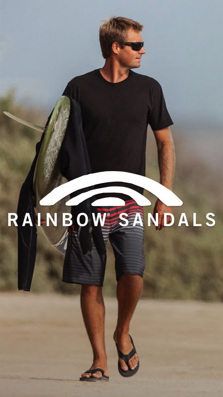 A man walking with a surfboard wearing flip-flops and casual clothing.