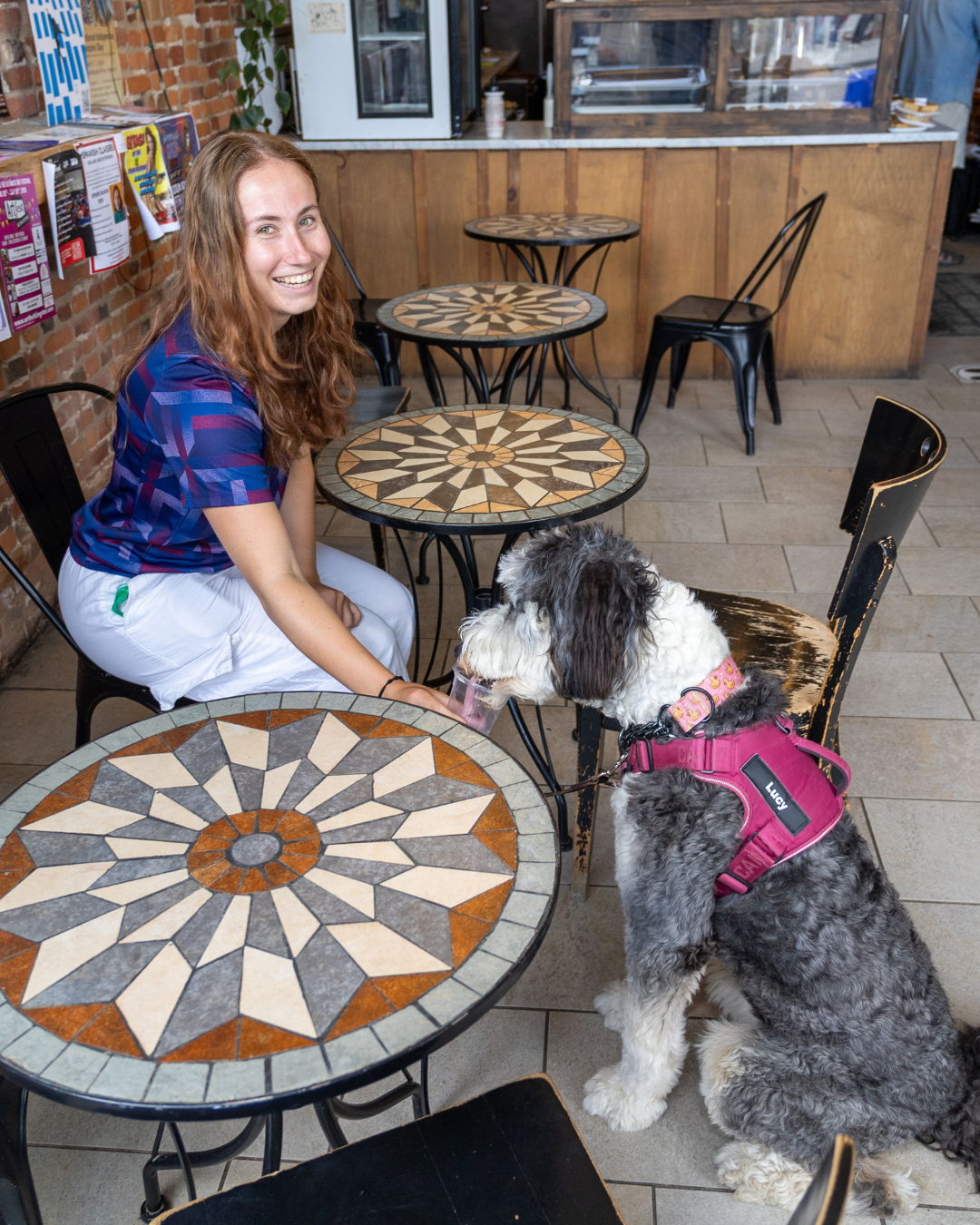A woman and a dog sit at mosaic-topped tables in a café.