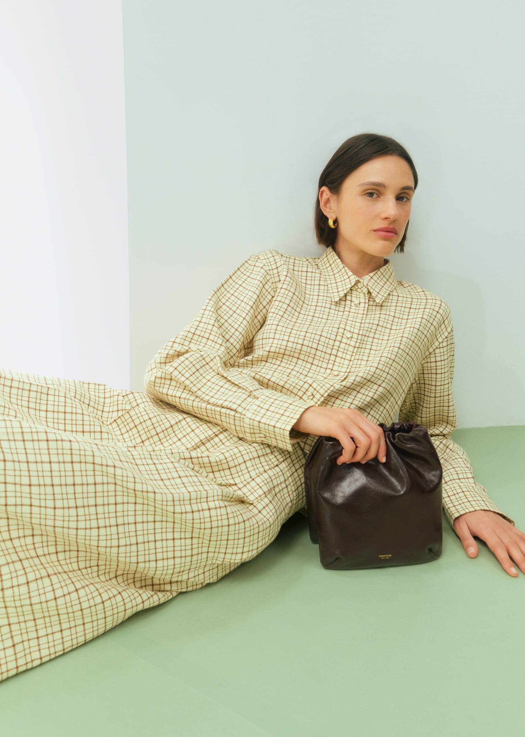 Woman in a patterned dress holding a bag, sitting on a pastel background.