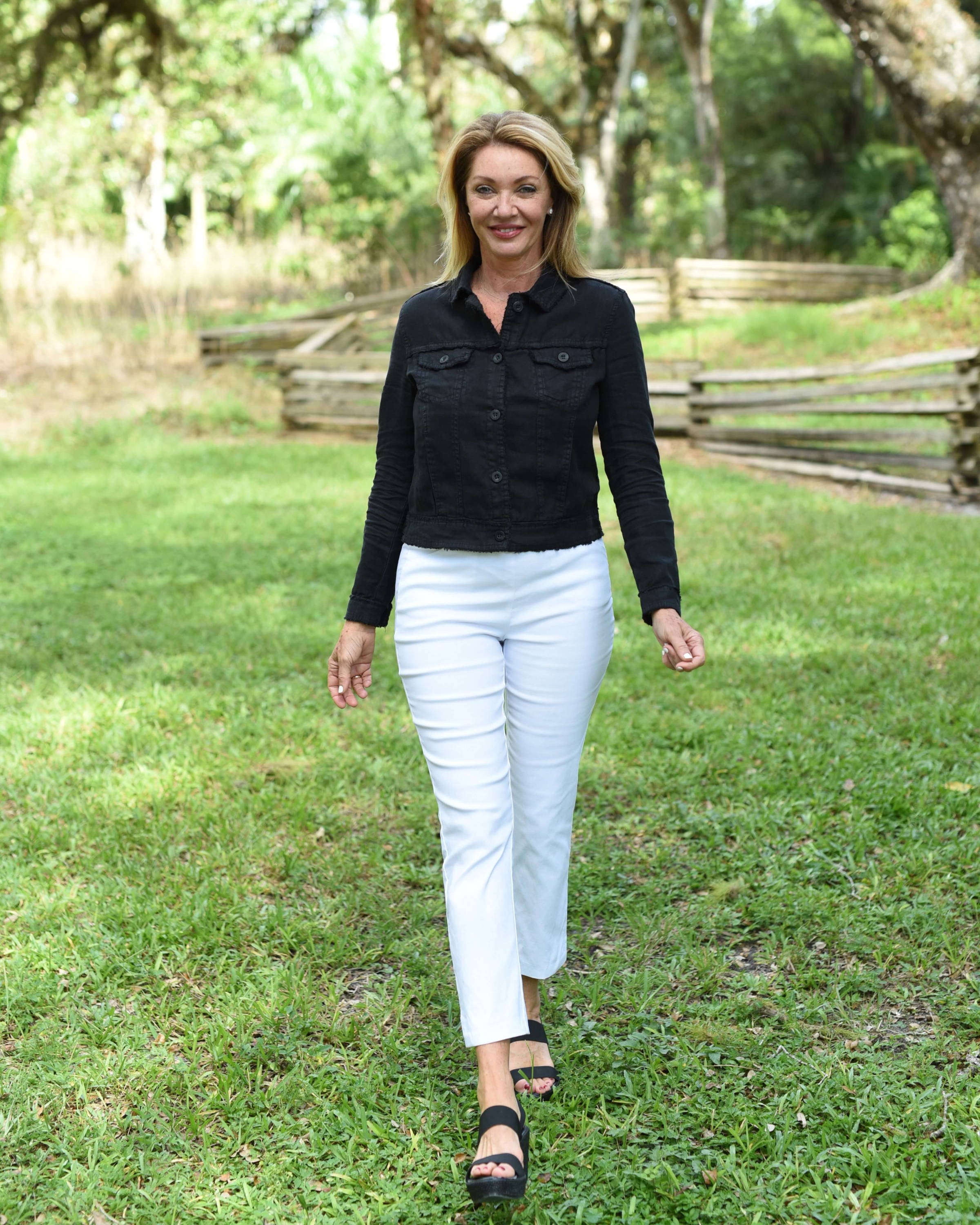 A blonde woman in a black jacket and white pants stands in a field with a tree in the background. 