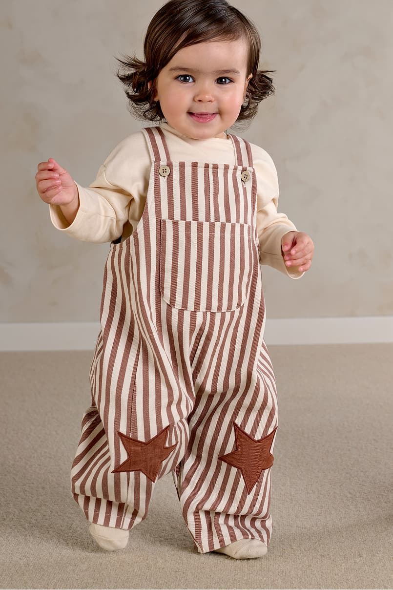 A smiling toddler in striped overalls with star patches.