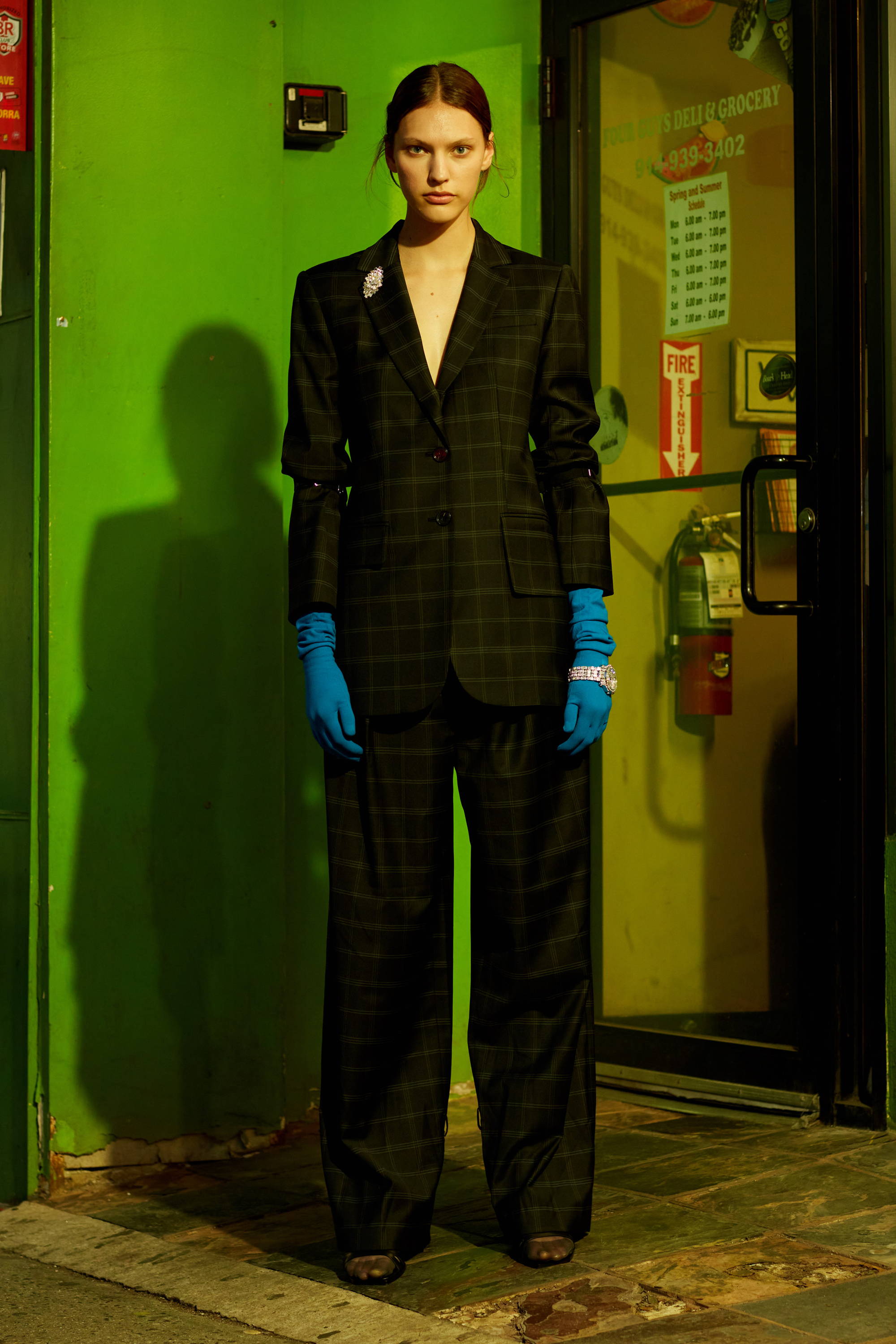 Model in a plaid suit with blue gloves, standing against a green wall.