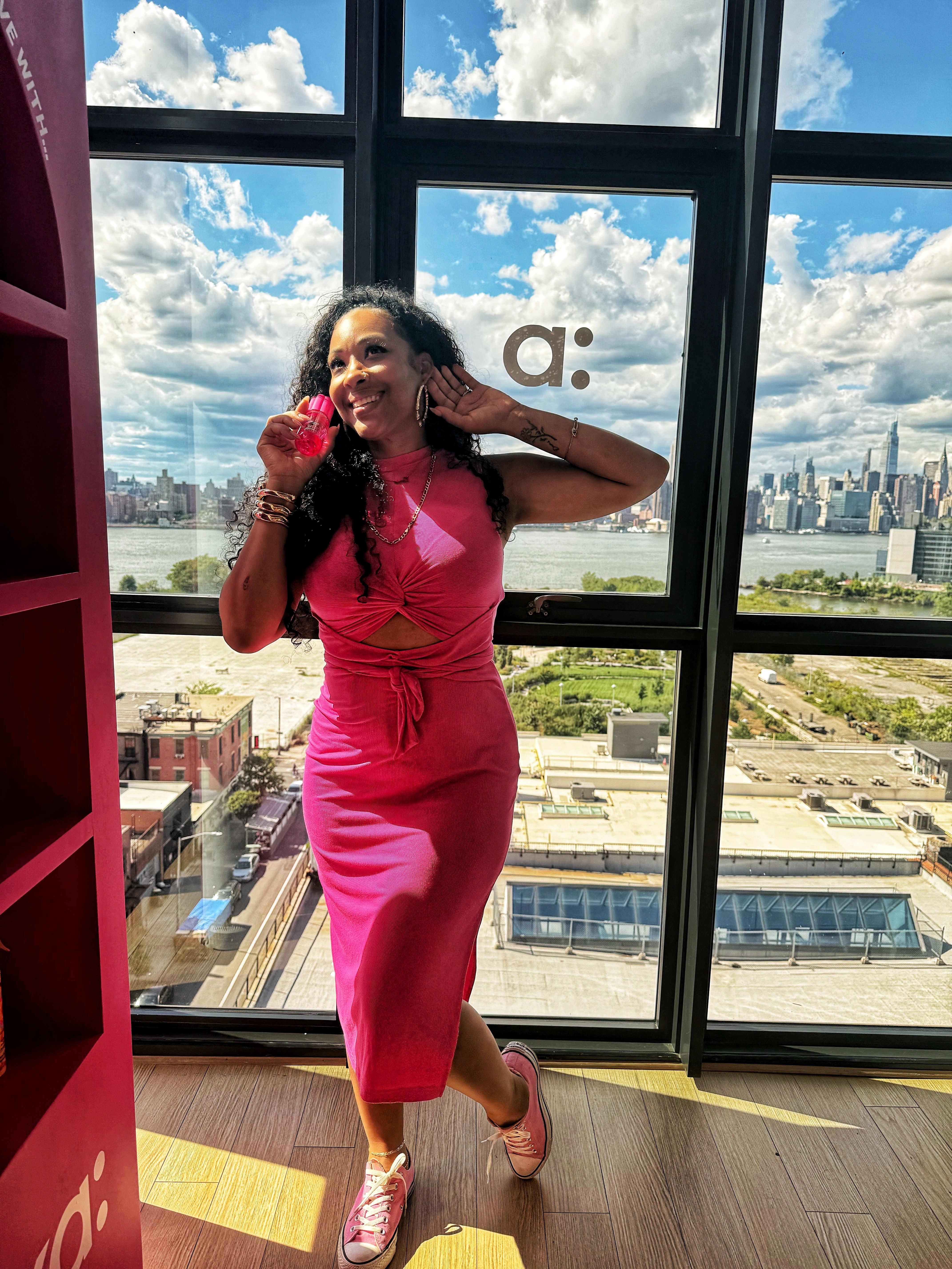 Woman in a pink dress posing near large windows with a city view.