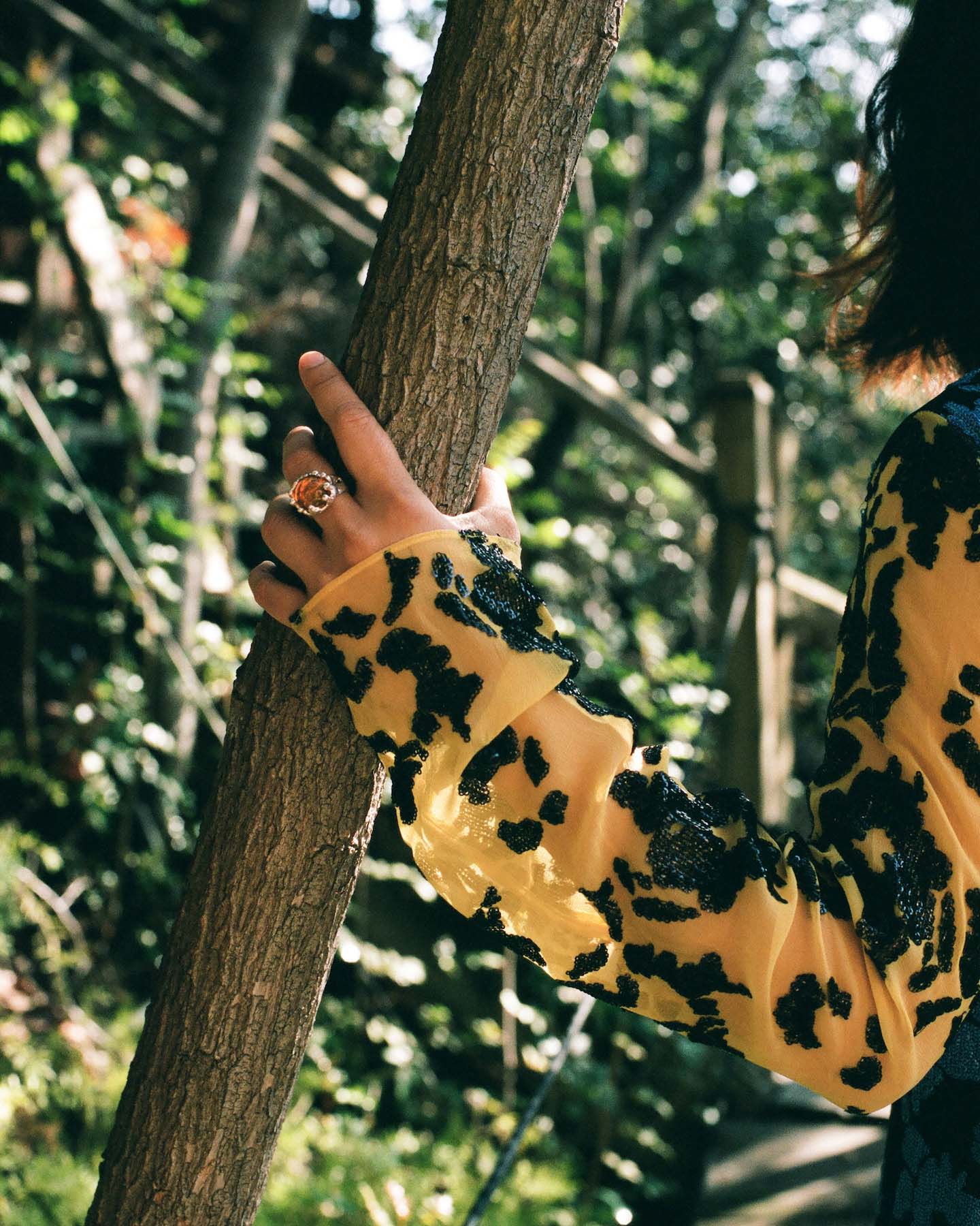A close up of a hand in a sequined and sheer dress and mondo mondo ring holding onto a tree.
