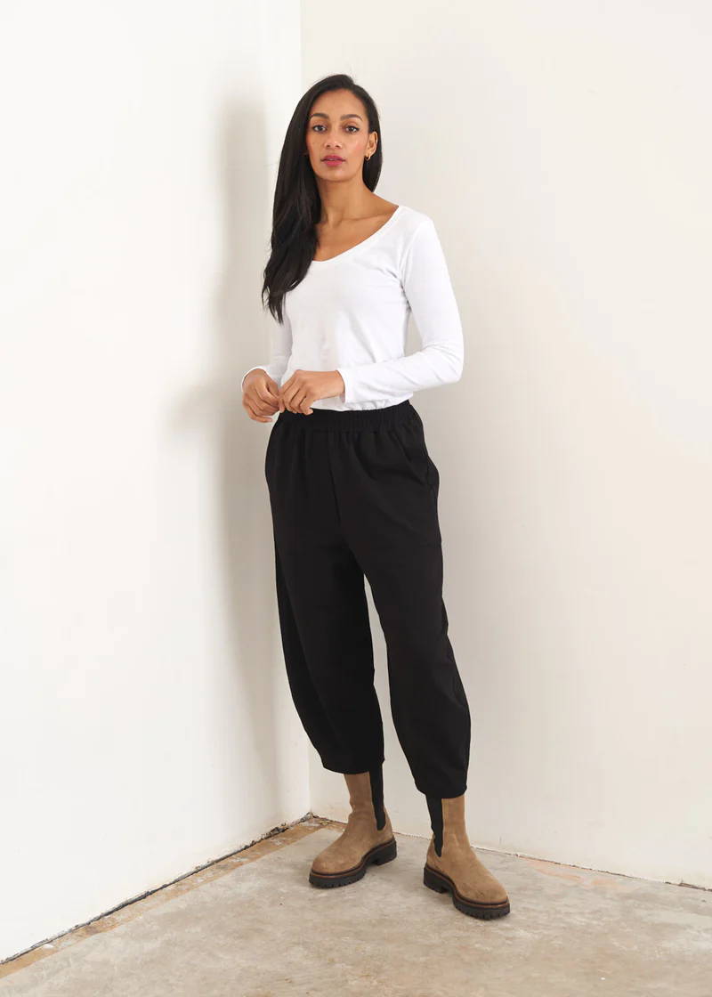 A model wearing a white long sleeved top weith black barrel leg jersey trousers and taupe coloured suede chelsea boots