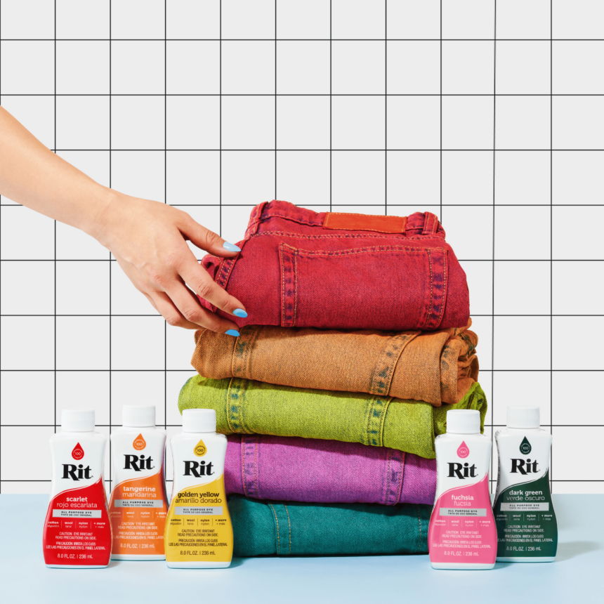 Hand reaching for stacked colorful jeans beside dye bottles on a grid background.