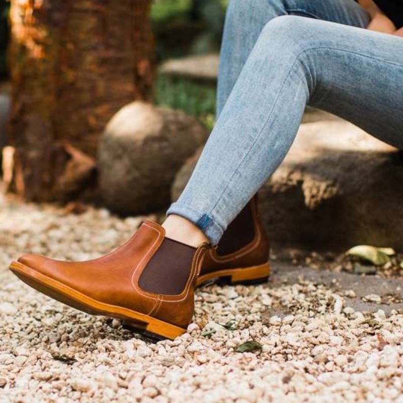 A side profile lifestyle view of a woman wearing the handcrafted Adelante Merida women's wide width leather Chelsea boots in a rich brown tone, sitting outdoors on a gravel ground.