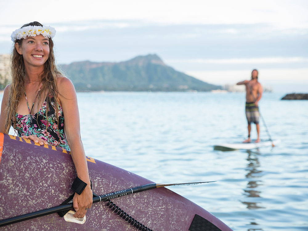 a woman holding a stand up paddleboard on a beach in hawaii with a guy paddleboarding in the background