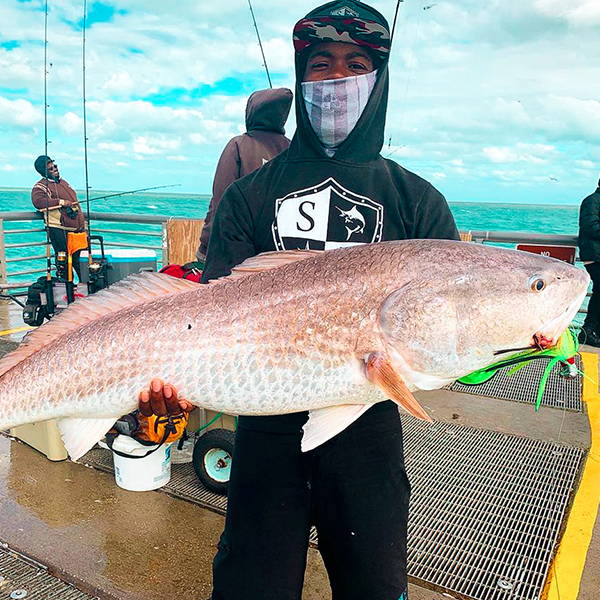 Borris Moore on a pier holding up a pink backed fish while they're wearing a black SA Company hoodie, a face shield over his face, and a SA Company hat with an camo underbrim design.
