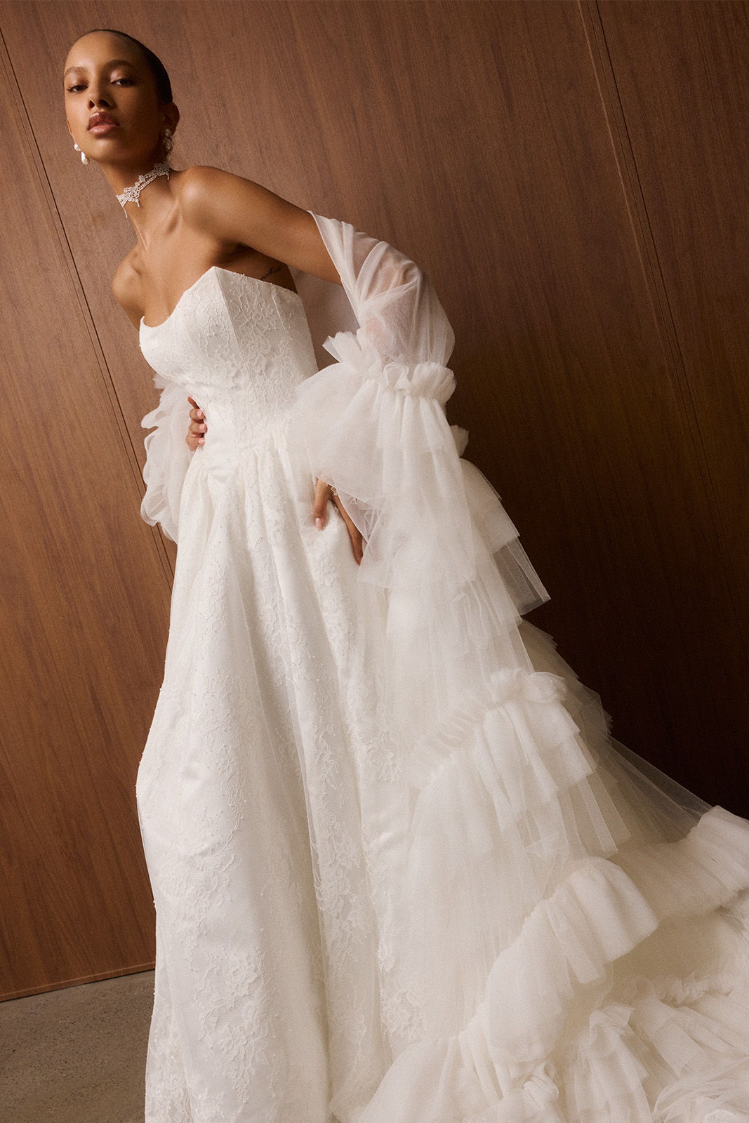 Model in a white wedding dress with ruffled details and sheer sleeves.