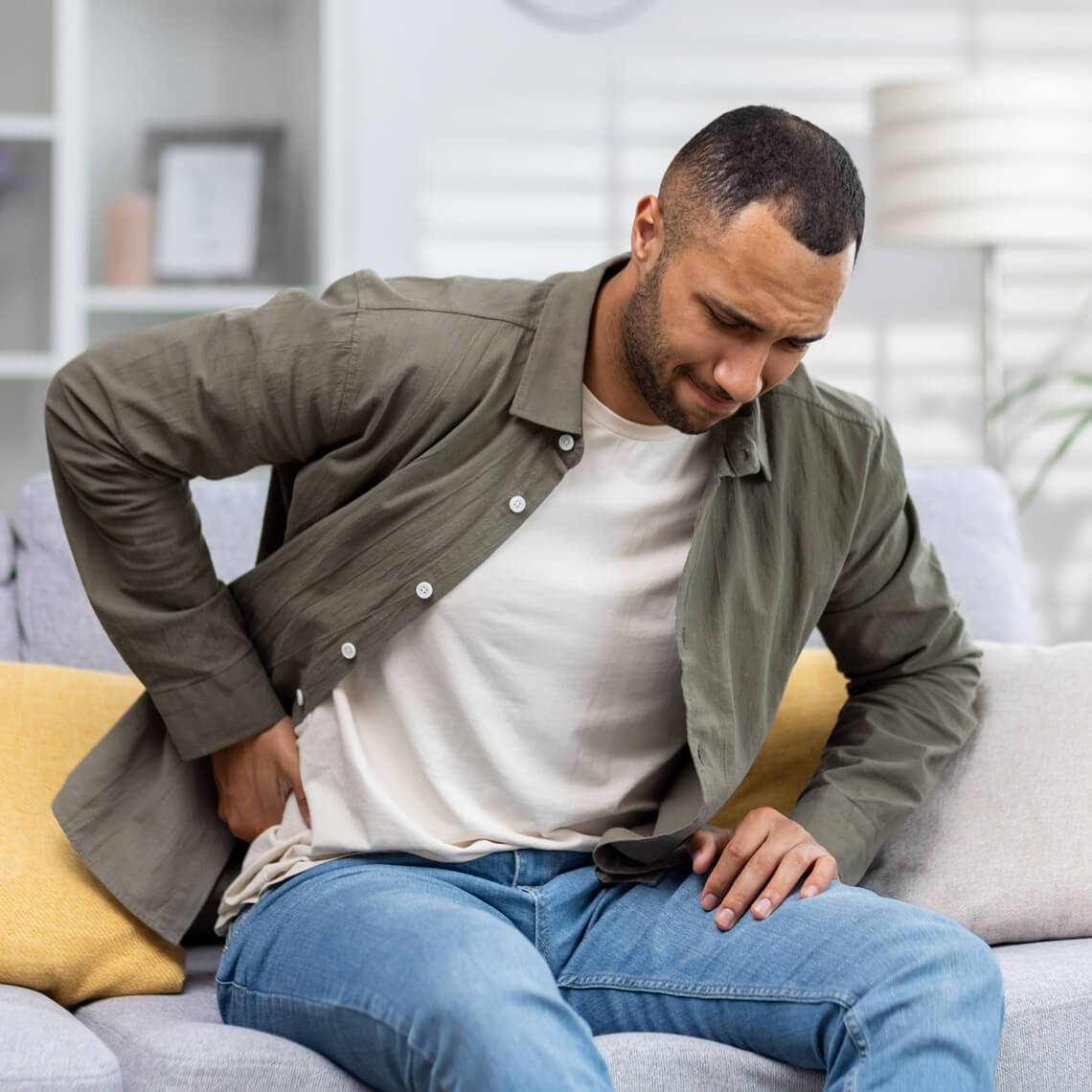 Man sitting on a couch, holding his lower back in discomfort.