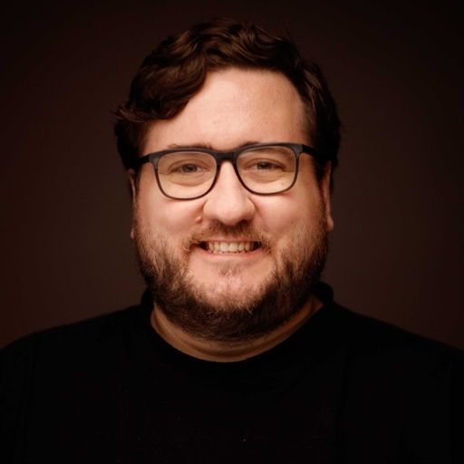 Smiling person with glasses and a beard against a dark background.