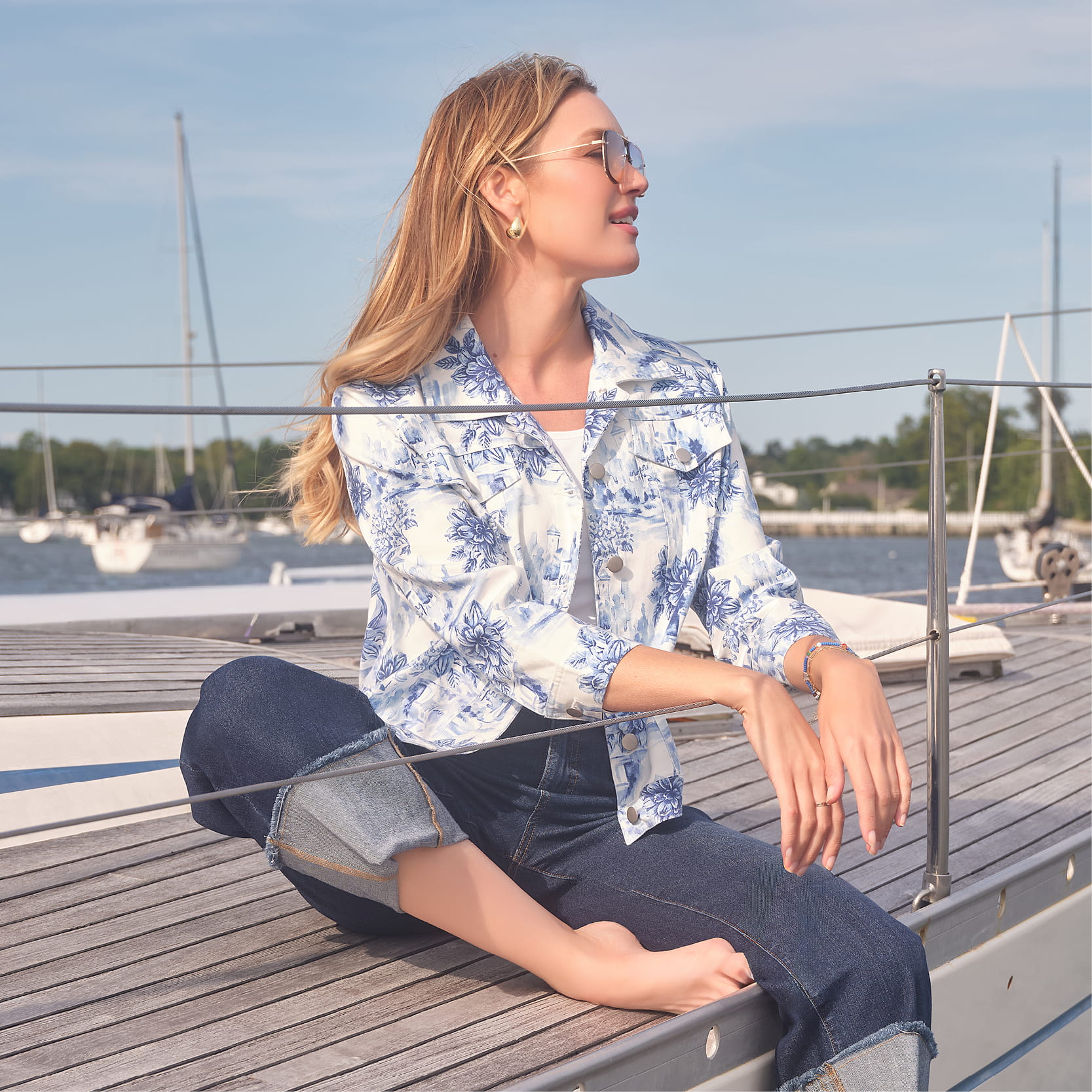 A woman sitting on a dock, wearing a floral shirt and jeans.