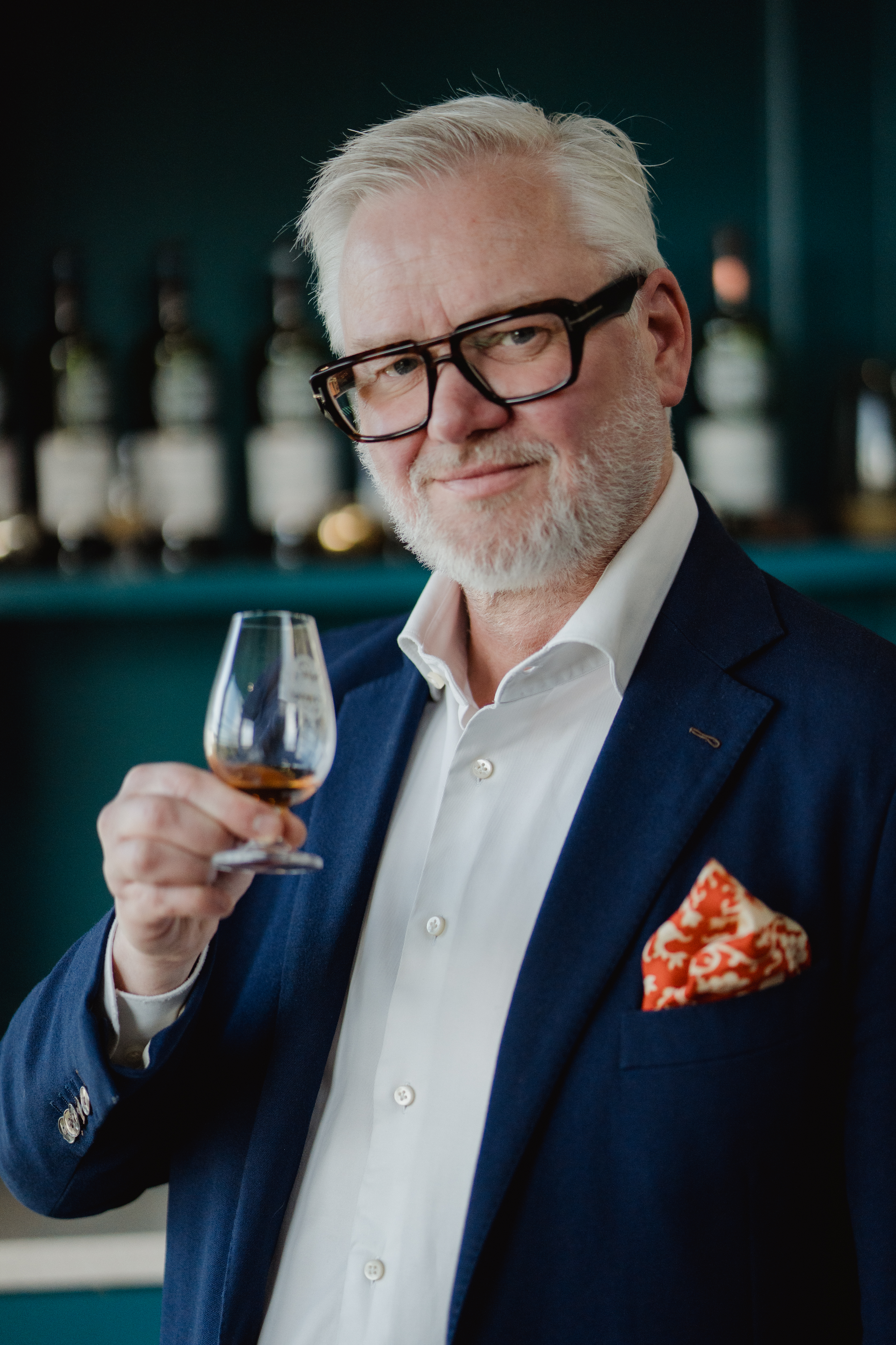 Man in a suit holding a glass of whiskey with bottles in background.