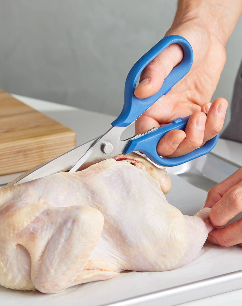 A chef using Blue Misen Kitchen Shears to remove the spine from a raw chicken.