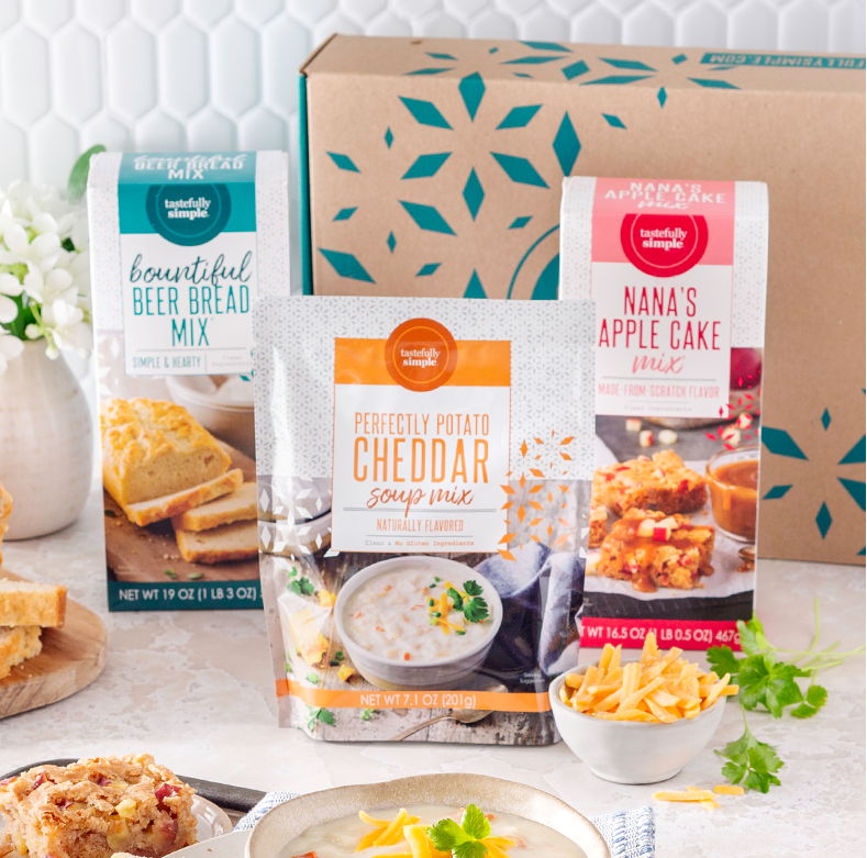 Bountiful Beer Bread Mix®, Perfectly Potato Cheddar Soup Mix and Nana’s Apple Cake Mix displayed with prepared foods and a Tastefully Simple gift box.