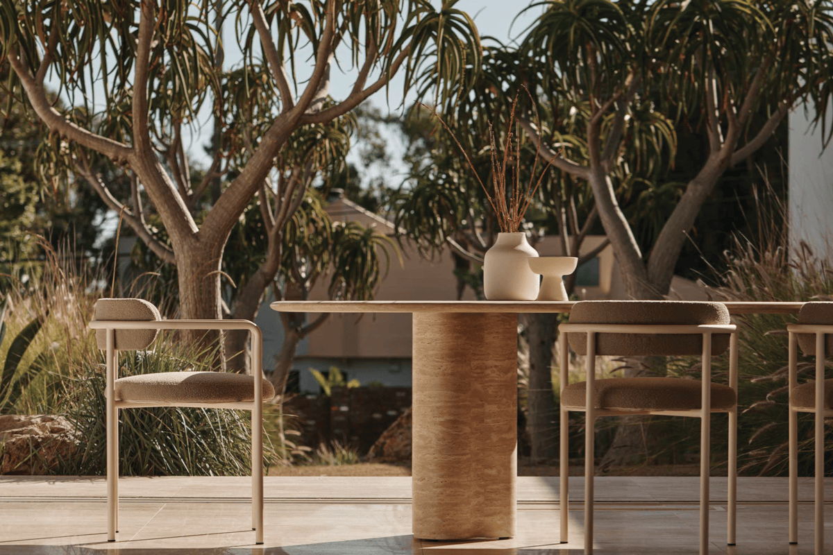 Outdoor dining setup with a travertine pedestal table, light framed dining chairs, and ceramic vessels, set against a desert garden view.