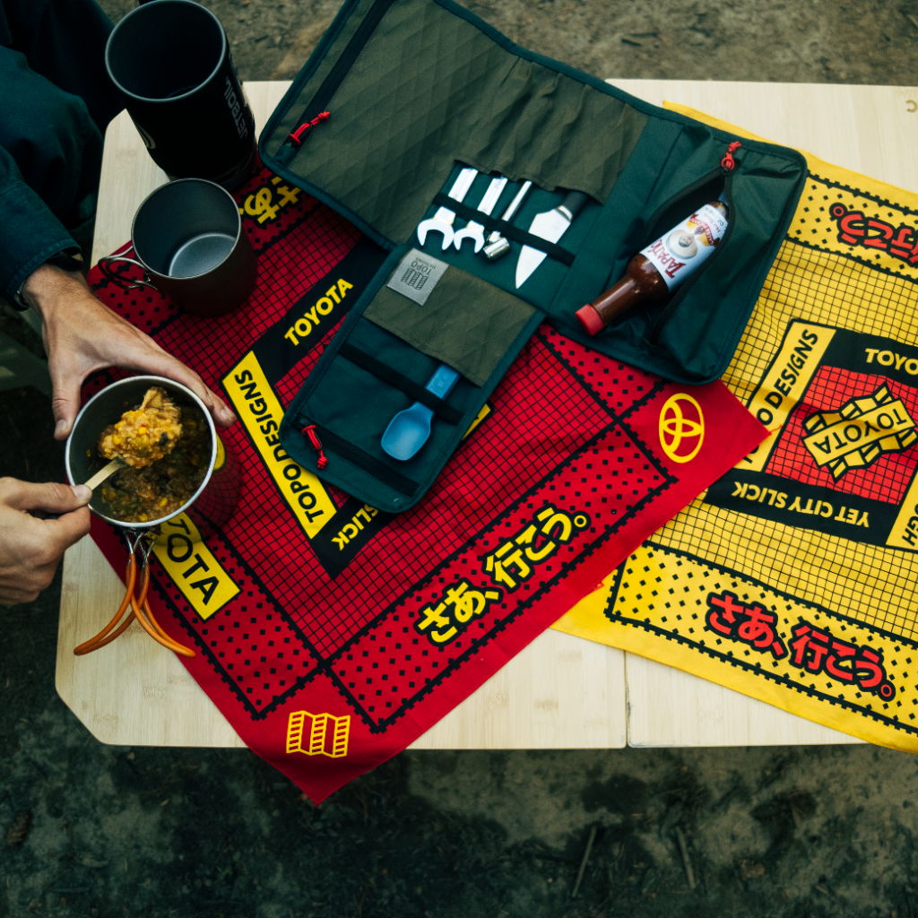 A person holding a bowl of noodles on colorful mats and camping gear.