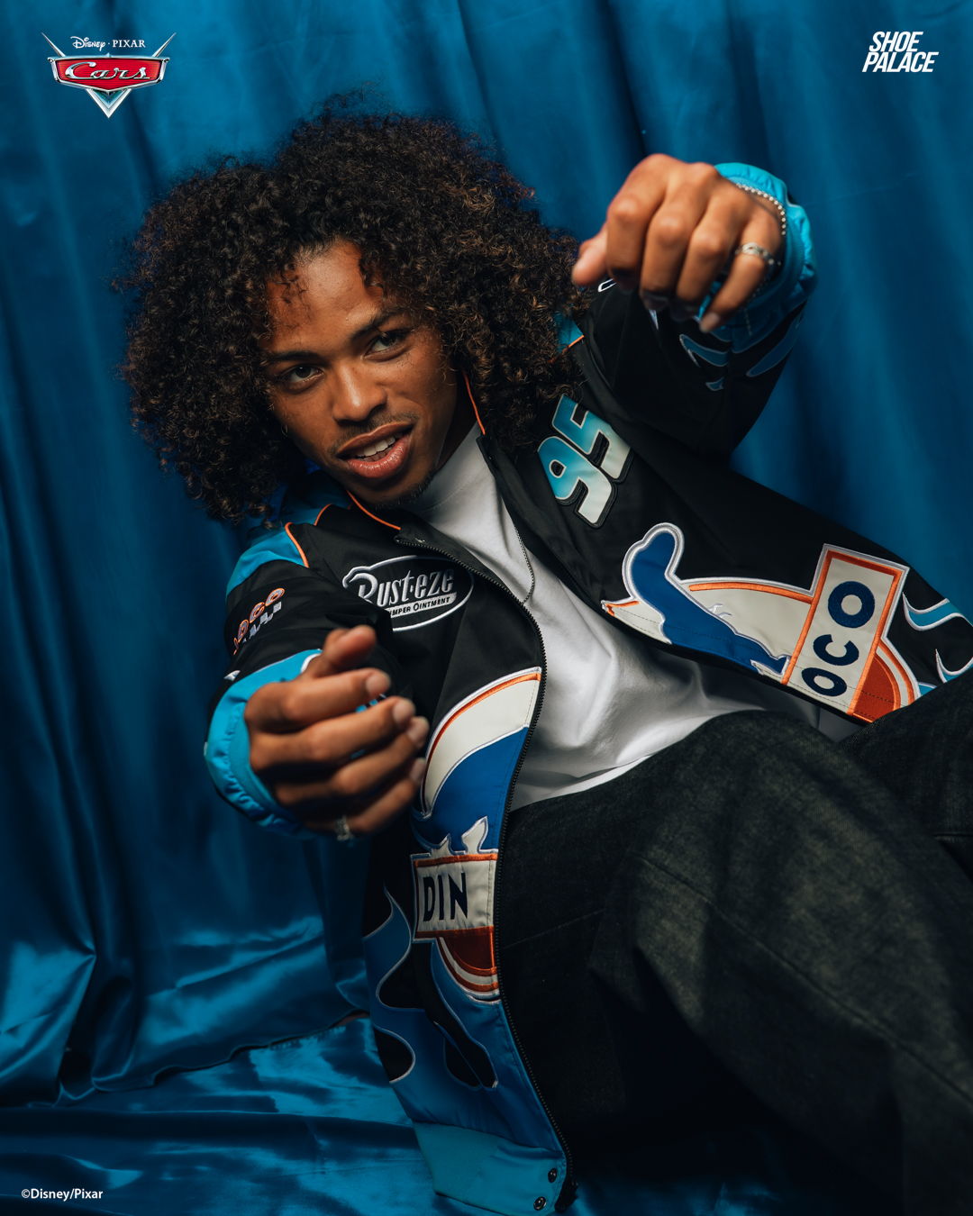 Person with curly hair in a colorful jacket posing against a blue backdrop.