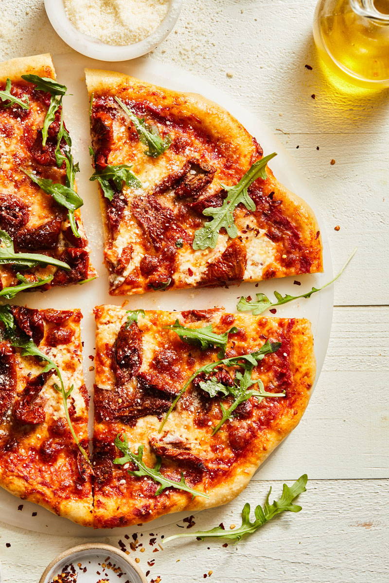 Cheesy baked pizza topped with sun-dried tomatoes and arugula and sliced for serving