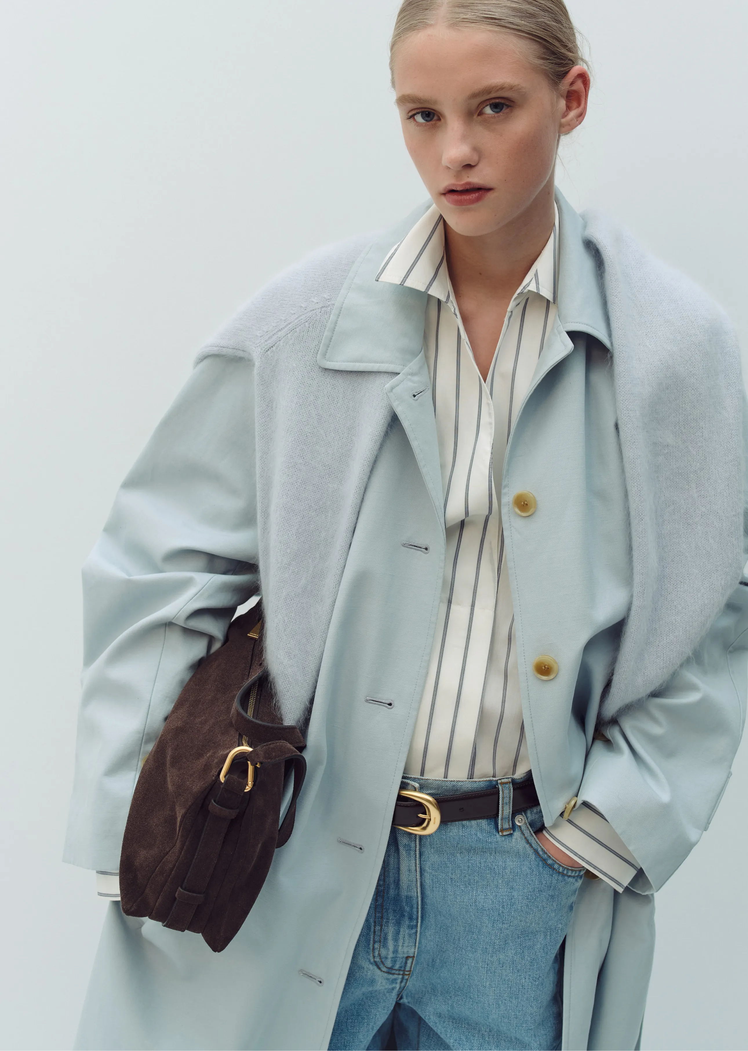 Model in light blue coat with brown bag, wearing striped shirt and jeans.