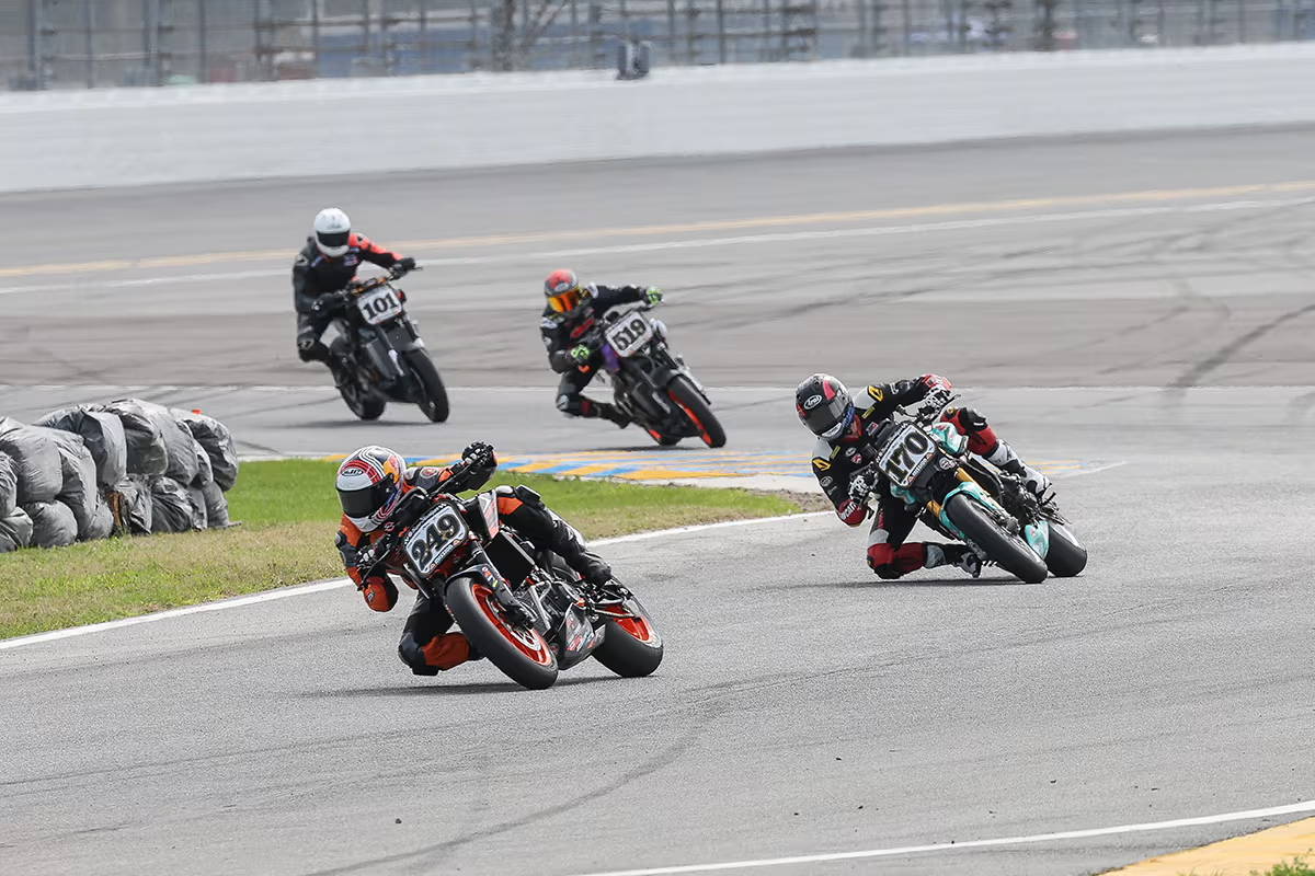 Motorcycle racers tightly grouped in a corner at Daytona International Speedway, showcasing intense racing action