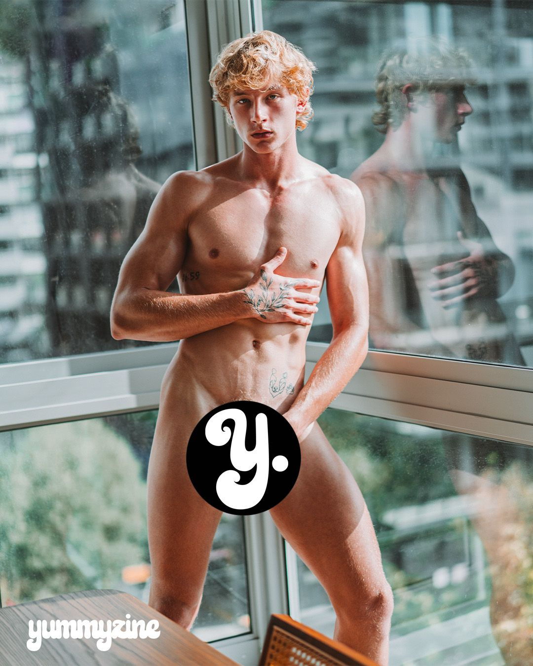 A man posing nude by a window with a city view.